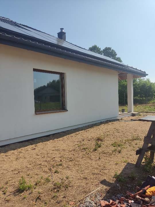 Boczny widok otynkowanego, parterowego domu z panelami słonecznymi na dachu i brązowymi ramami okiennymi, z widocznym fragmentem terenu przed budynkiem.