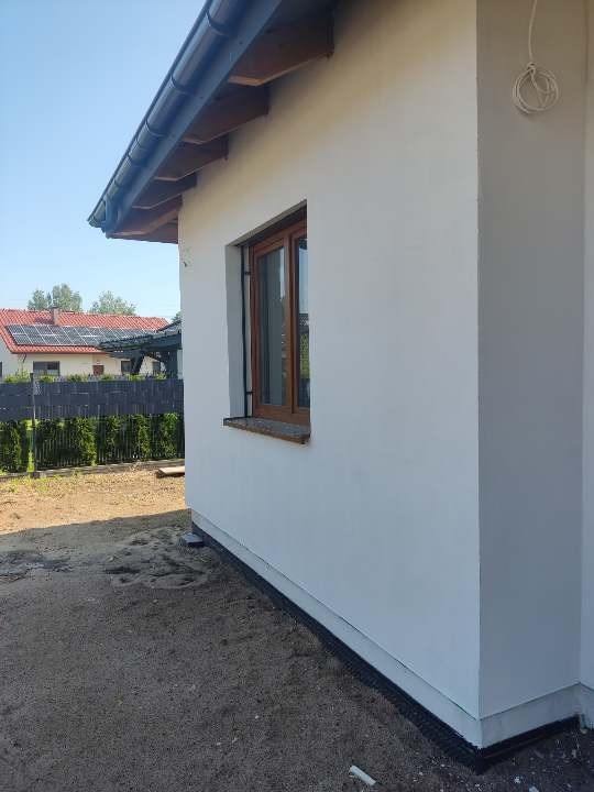 Świeżo otynkowana ściana domu jednorodzinnego z brązowym oknem, widoczne przewody elektryczne na rogu budynku, w tle dom z panelami słonecznymi.