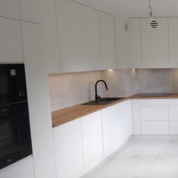 eMCe- Studio - Nowoczesna kuchnia z białymi frontami, drewnianym blatem i czarną armaturą, oświetlenie LED pod szafkami wiszącymi, widoczny piekarnik w zabudowie.