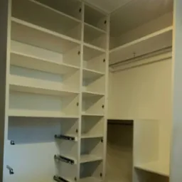 Biała szafa wnękowa z otwartymi półkami i drążkami na ubrania, widoczne szuflady z metalowymi prowadnicami, drewniana podłoga.