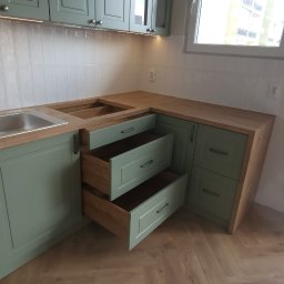 eMCe- Studio - Fragment kuchni z otwartymi szufladami w odcieniach zieleni i drewna, widoczne płytki na ścianie i parkiet w jodełkę na podłodze.