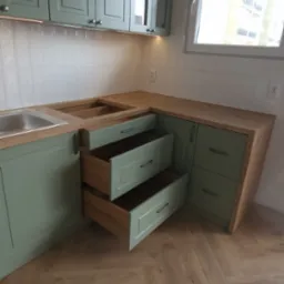 Fragment kuchni z otwartymi szufladami w odcieniach zieleni i drewna, widoczne płytki na ścianie i parkiet w jodełkę na podłodze.