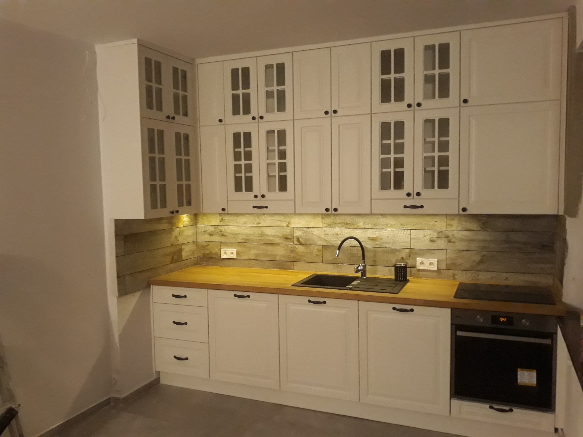 Biała zabudowa kuchenna z drewnianym blatem i ścianą wykończoną panelami imitującymi drewno, widoczny piekarnik i czarny zlew.