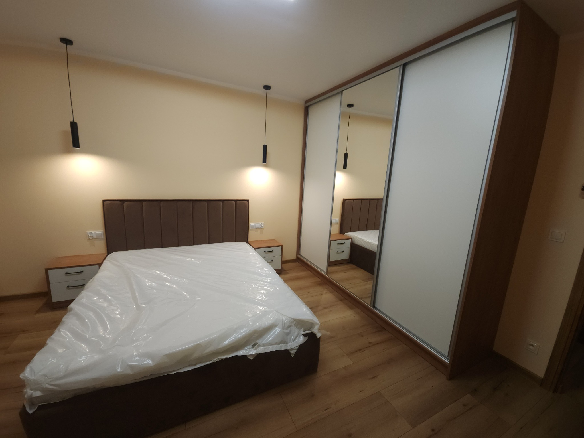 Nowoczesna sypialnia z łóżkiem z brązowym zagłówkiem i szafą wnękową z przesuwnymi drzwiami, częściowo lustrzanymi, podwieszane lampy punktowe.