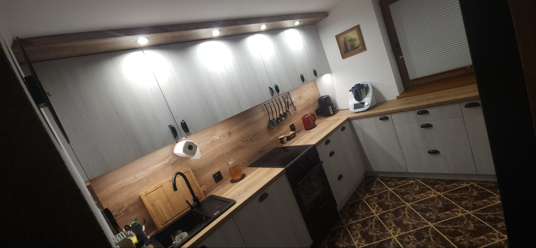 Nowoczesna kuchnia z szarymi szafkami, drewnianym blatem i brązowymi płytkami podłogowymi. Widoczne akcesoria kuchenne i oświetlenie punktowe.