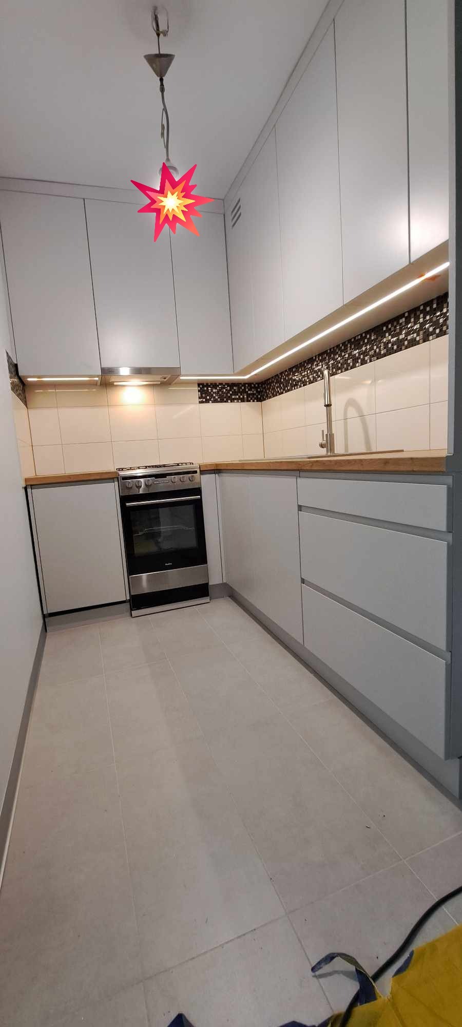 Nowoczesna kuchnia w odcieniach szarości z drewnianym blatem, oświetleniem LED pod szafkami i oryginalną lampą w kształcie gwiazdy.