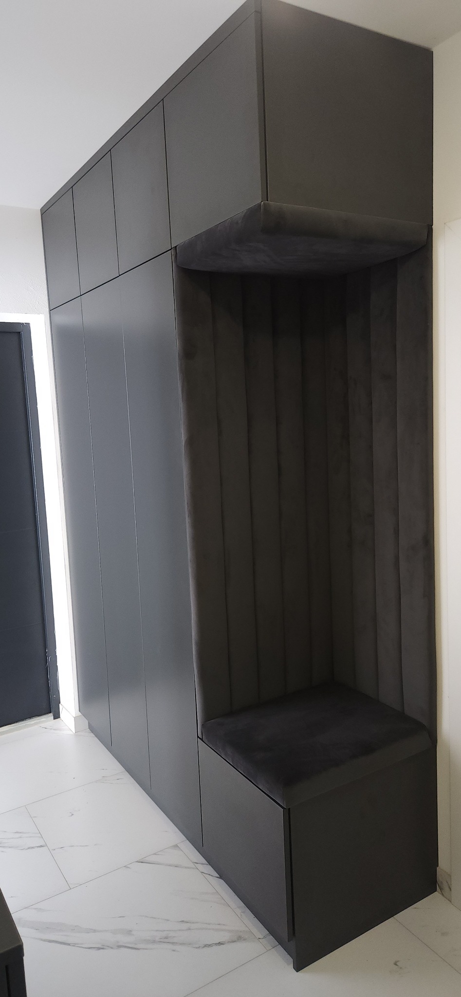 Ciemnoszara szafa w przedpokoju z siedziskiem obitym ciemnym welurem, wbudowana wnęka z tapicerowanym oparciem w pionowe pasy, marmurowe płytki na podłodze.