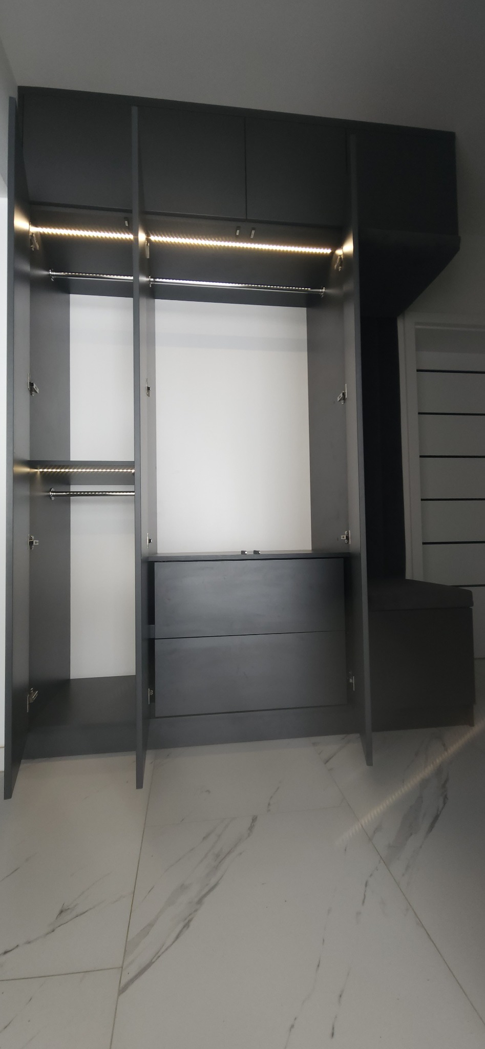 Szara szafa wnękowa z oświetleniem LED i otwartymi drzwiami, ukazująca półki i szuflady, na tle białej podłogi z marmurowym wzorem.