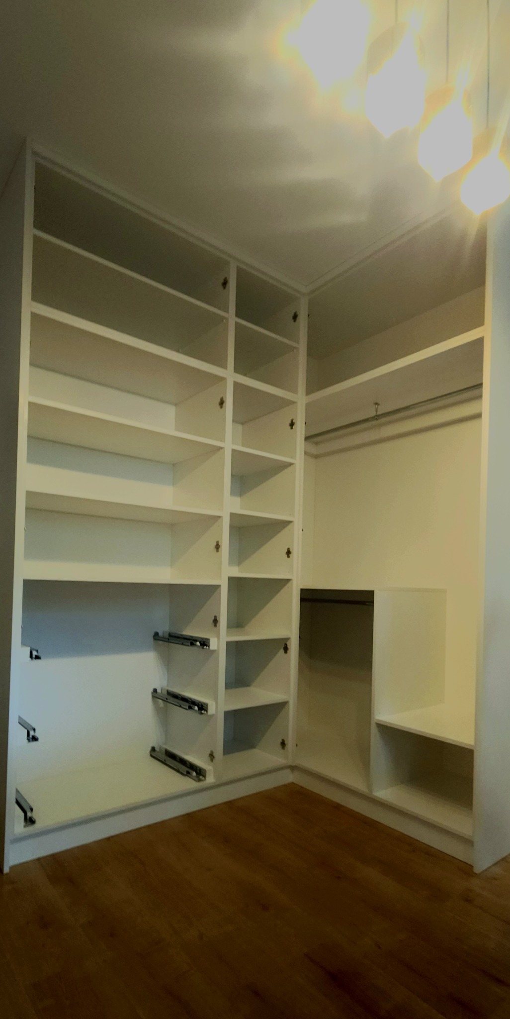 Biała szafa wnękowa z otwartymi półkami i drążkami na ubrania, widoczne szuflady z metalowymi prowadnicami, drewniana podłoga.