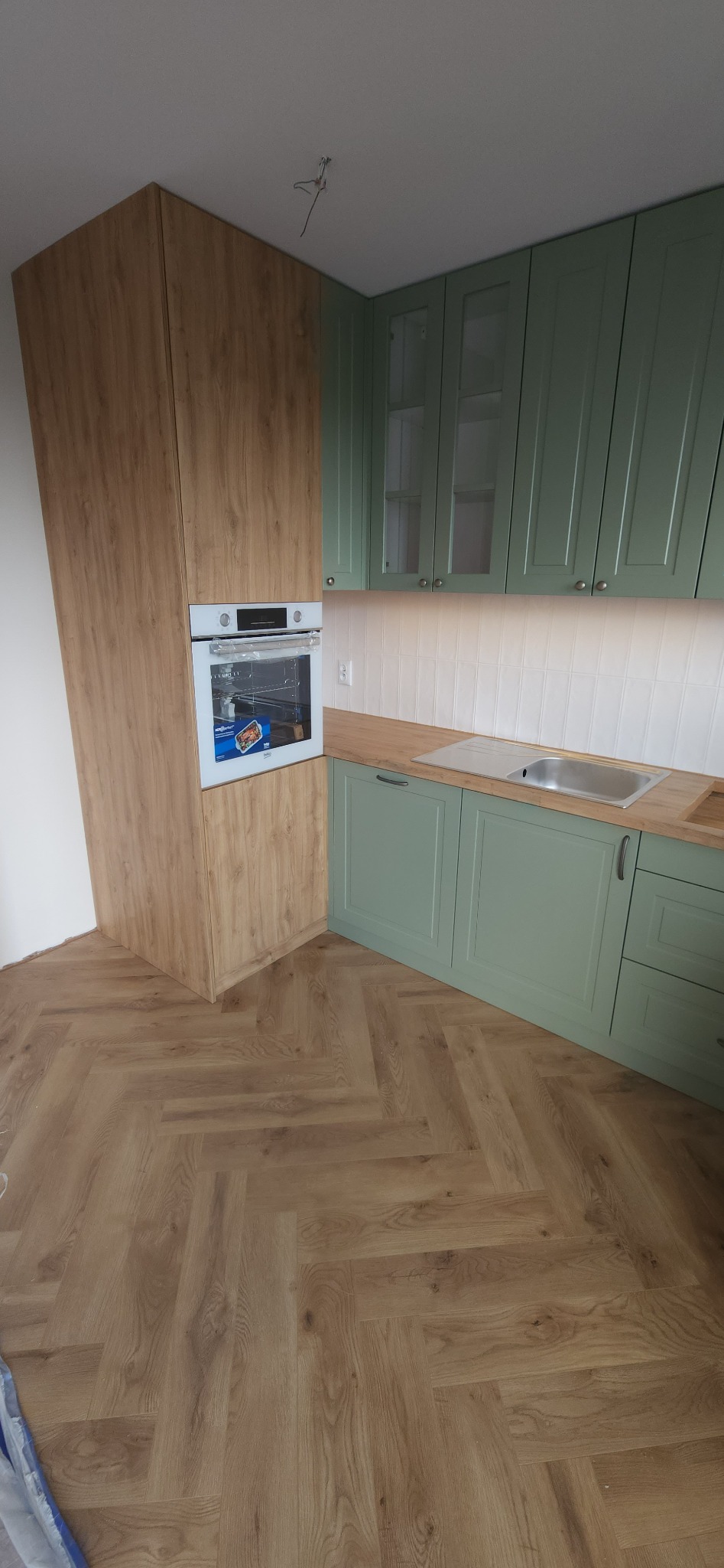 Kuchenna zabudowa z zielonymi szafkami i drewnianym blatem, piekarnik wkomponowany w wysoką szafkę z drewnopodobnym frontem, parkiet ułożony w jodełkę.