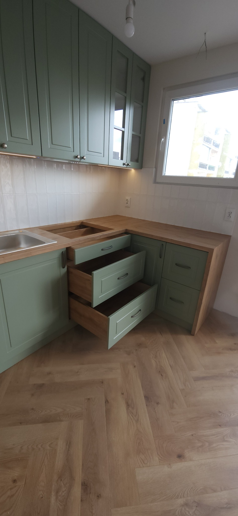 Fragment kuchni z otwartymi szufladami w odcieniach zieleni i drewna, widoczne płytki na ścianie i parkiet w jodełkę na podłodze.