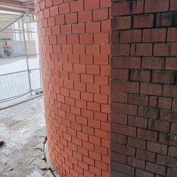 E-M Jerzy MIOSGA - Cylindryczna ściana z czerwonej cegły, częściowo wyczyszczona, kontrastująca z brudną, ciemniejszą częścią, widoczne rusztowania w tle.