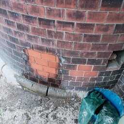 E-M Jerzy MIOSGA - Fragment okrągłej, ceglanej elewacji z widocznym obszarem po czyszczeniu, obok niebieskie wiadro z zielonymi rękawicami ochronnymi.