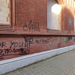 E-M Jerzy MIOSGA - Ceglana elewacja budynku z widocznymi graffiti i częściowo zamurowanymi oknami, sfotografowana z poziomu chodnika z kostki brukowej.