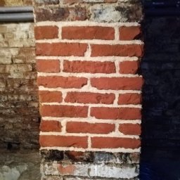 E-M Jerzy MIOSGA - Ceglany filar z widocznymi śladami zabrudzeń i miejscami pokrytymi mchem, sąsiadujący z ciemną ścianą i fragmentem ściany z rurami.