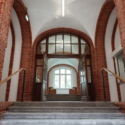 E-M Jerzy MIOSGA - Wnętrze budynku z ceglanymi łukami, schodami i otwartymi drzwiami, widok z dołu na jasne okno w tle.