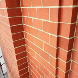 Czerwona ceglana elewacja narożna z jasną fugą, widoczna z dołu, z fragmentem drabiny w lewym dolnym rogu.