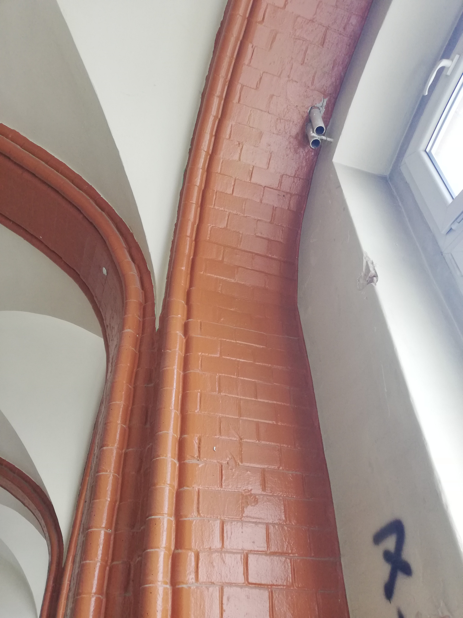 Ceglany łuk architektoniczny wewnątrz budynku, z widoczną instalacją elektryczną i fragmentem okna.