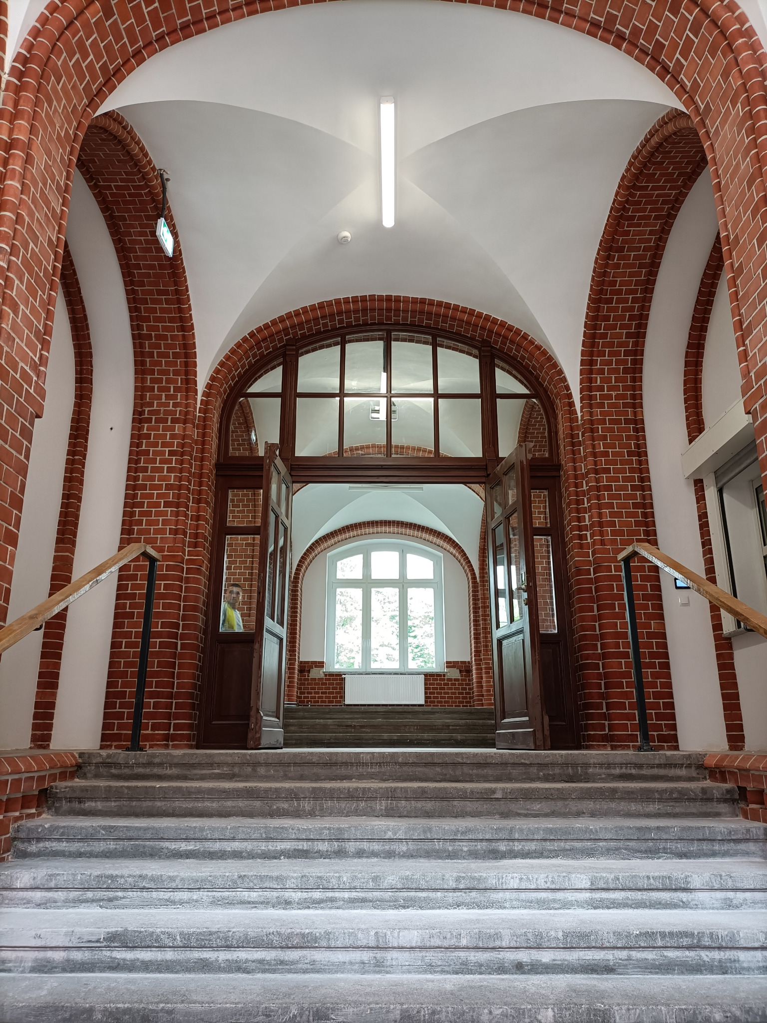 Wnętrze budynku z ceglanymi łukami, schodami i otwartymi drzwiami, widok z dołu na jasne okno w tle.