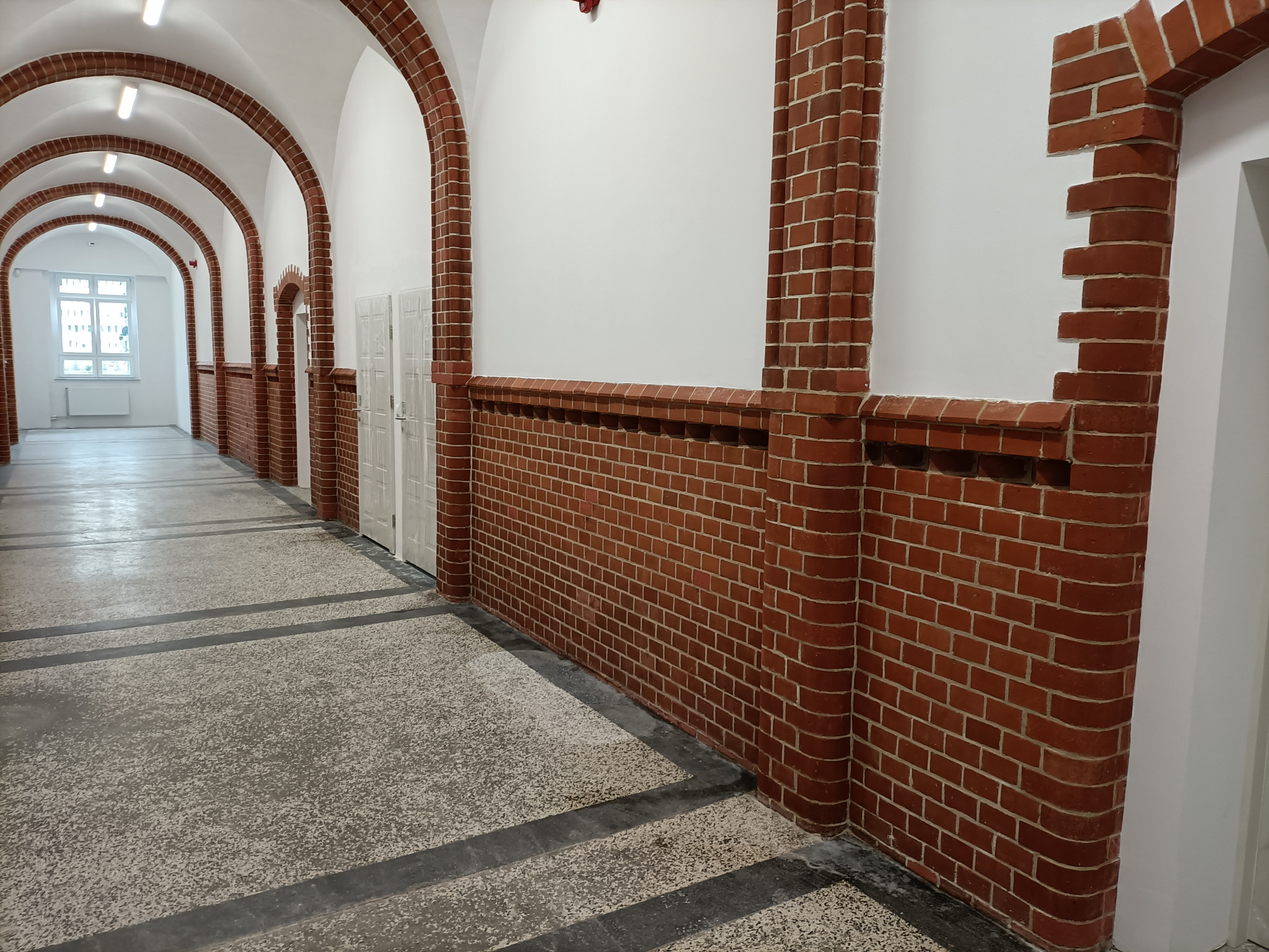 Długi korytarz z ceglanymi łukami i ścianami, widok w perspektywie, szaro-biała posadzka z ciemnymi pasami.