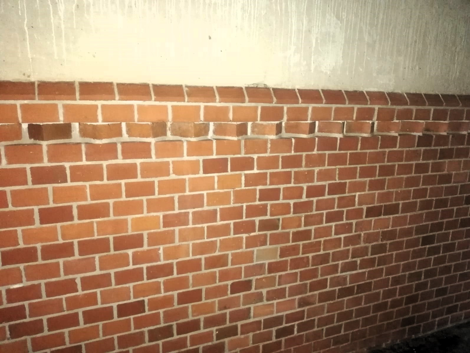 Ceglana ściana z wystającymi cegłami tworzącymi dekoracyjny wzór, widoczne ślady zacieków na górnej części ściany.