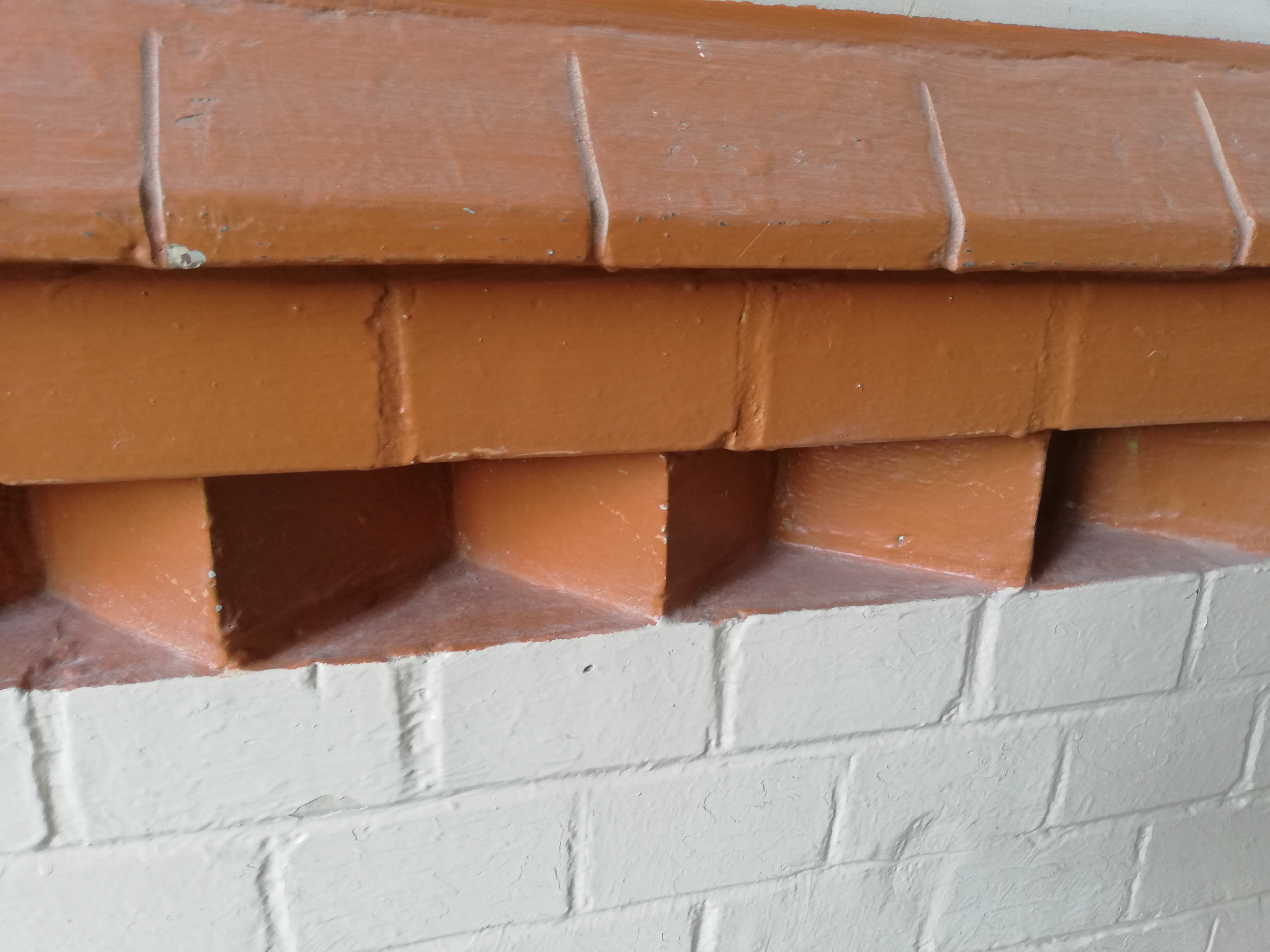 Pomarańczowa, pomalowana ceglana elewacja z dekoracyjnymi wgłębieniami, kontrastująca z białą, pomalowaną cegłą poniżej.