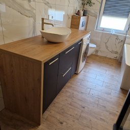 KSmeble Kamil Sławik - Łazienka z umywalką nablatową w kształcie łzy, złotą armaturą i szafką z drewnianym blatem i grafitowymi frontami, obok pralka.