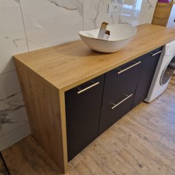 KSmeble Kamil Sławik - Łazienka z szafką na wymiar z blatem imitującym drewno, umywalką nablatową w kształcie łódki z złotym rantem, ciemnymi frontami szuflad z podłużnymi uchwytami, pralką i toaletą w tle.