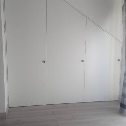 KSmeble Kamil Sławik - Biała szafa wnękowa z trzema drzwiami, dopasowana do skosu dachu, z szarymi okrągłymi uchwytami na tle szarej podłogi z paneli i fragmentu szarego pufa.