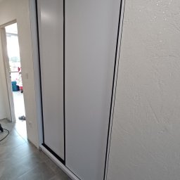 KSmeble Kamil Sławik - Biała szafa wnękowa z przesuwnymi drzwiami, czarne obramowanie, szare płytki podłogowe, ściana z dekoracyjnym tynkiem.