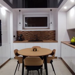 KSmeble Kamil Sławik - Nowoczesna kuchnia z białymi szafkami, drewnianym blatem i mozaiką, okrągły stół z czterema krzesłami w centrum, czarne, lśniące wykończenie sufitu.