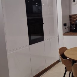 KSmeble Kamil Sławik - Biała, minimalistyczna zabudowa kuchenna z wbudowanym piekarnikiem i mikrofalówką Amica, widoczny fragment drewnianego stołu i krzesła w tle.