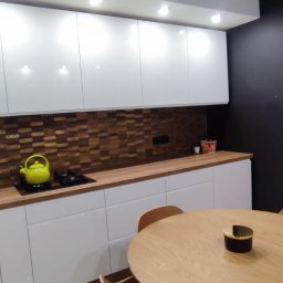 KSmeble Kamil Sławik - Nowoczesna kuchnia z białymi, błyszczącymi szafkami, drewnianym blatem i mozaiką na ścianie. Na blacie stoi zielony czajnik obok płyty indukcyjnej. W tle okrągły stół z drewna.