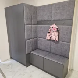 Szara zabudowa przedpokoju z szafą, siedziskiem i panelem tapicerowanym, z zawieszoną kurtką dziecięcą w kwiaty na wieszaku, widoczne fragmenty drabiny i podłogi z płytek imitujących marmur.