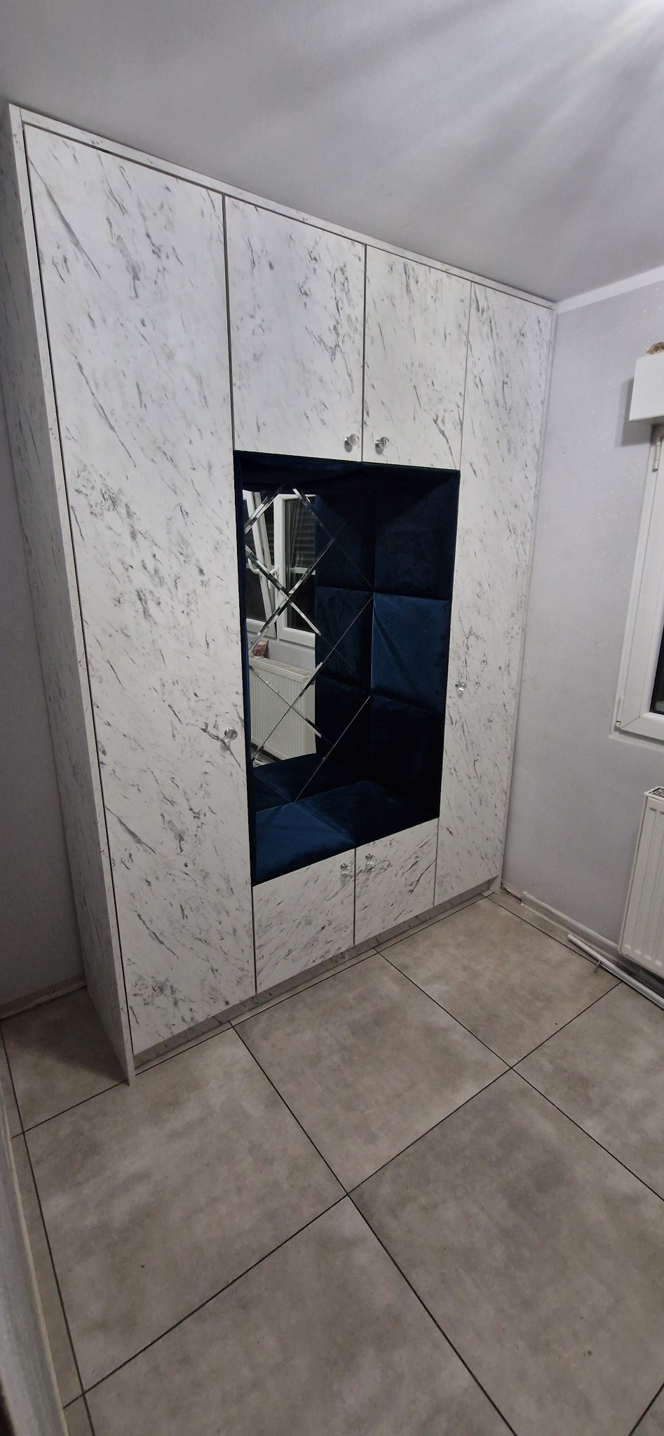 Biała szafa z marmurowym wzorem, centralny element z lustrami i niebieskim tapicerowanym wnętrzem, kryształowe gałki, szara podłoga.