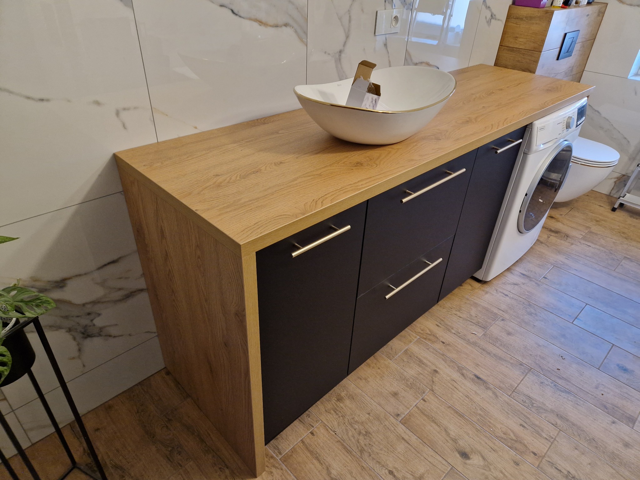 Łazienka z szafką na wymiar z blatem imitującym drewno, umywalką nablatową w kształcie łódki z złotym rantem, ciemnymi frontami szuflad z podłużnymi uchwytami, pralką i toaletą w tle.