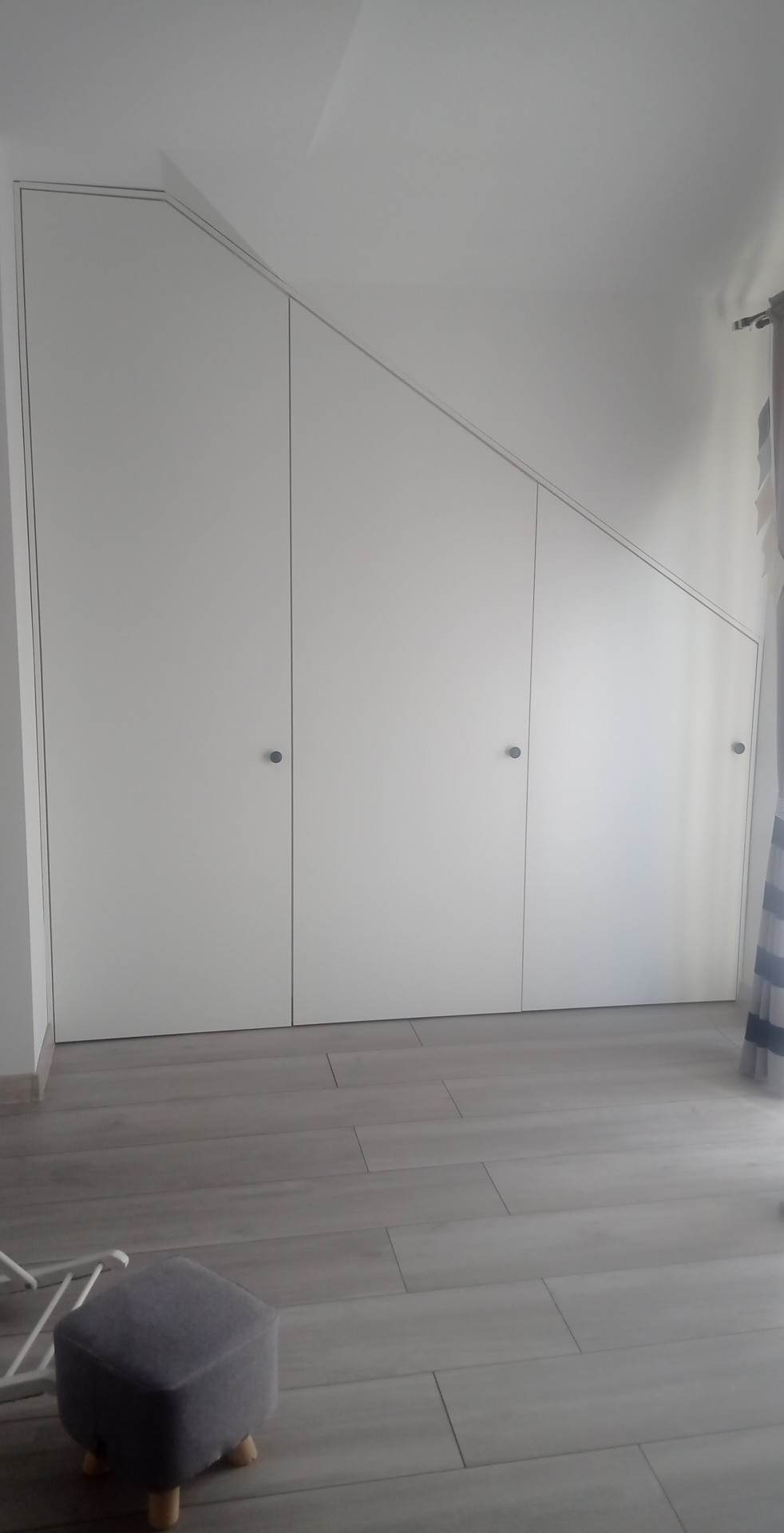 Biała szafa wnękowa z trzema drzwiami, dopasowana do skosu dachu, z szarymi okrągłymi uchwytami na tle szarej podłogi z paneli i fragmentu szarego pufa.