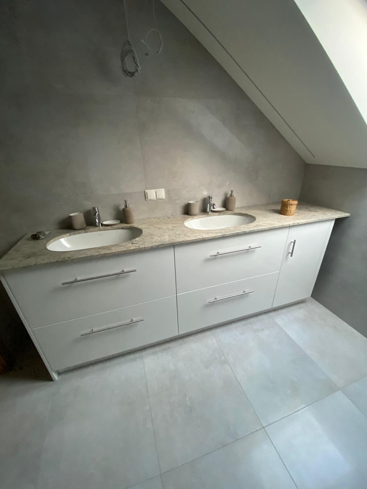 Biała szafka łazienkowa z dwoma umywalkami i blatem z jasnego kamienia w łazience na poddaszu z szarą glazurą.
