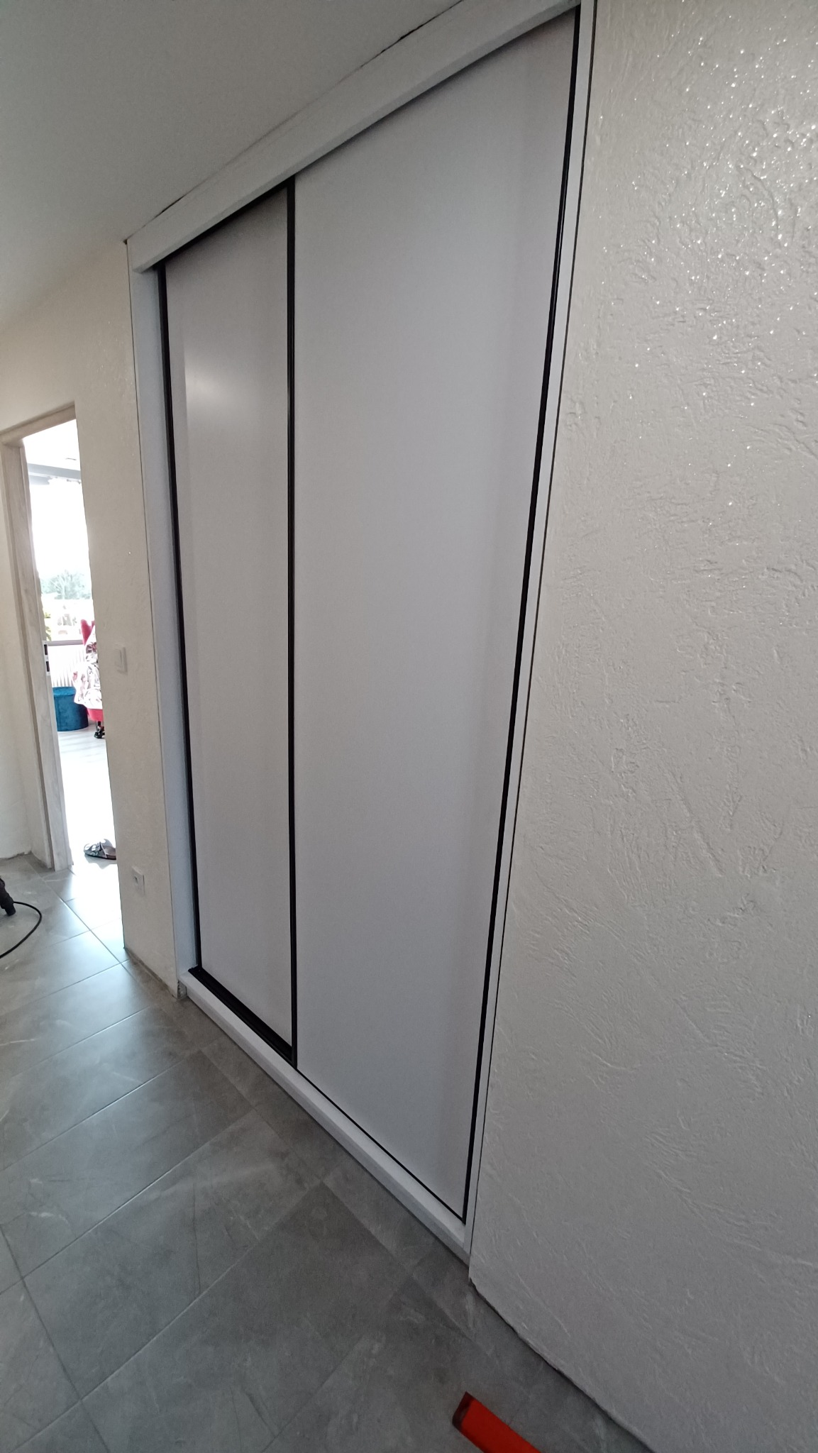 Biała szafa wnękowa z przesuwnymi drzwiami, czarne obramowanie, szare płytki podłogowe, ściana z dekoracyjnym tynkiem.