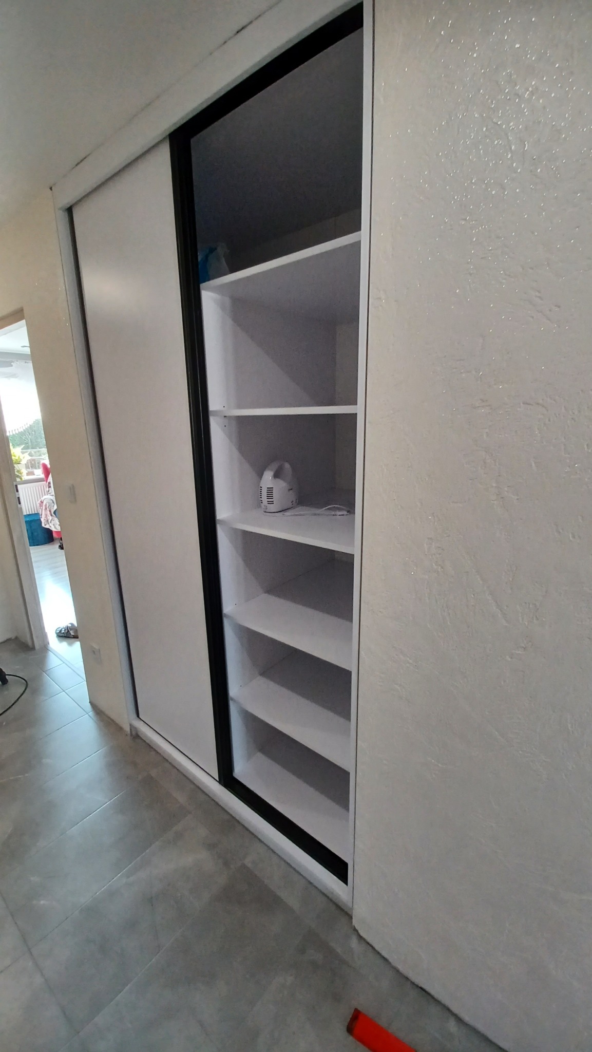 Biała szafa wnękowa z przesuwnymi drzwiami, czarną ramą i pustymi półkami, na jednej z półek stoi biały inhalator