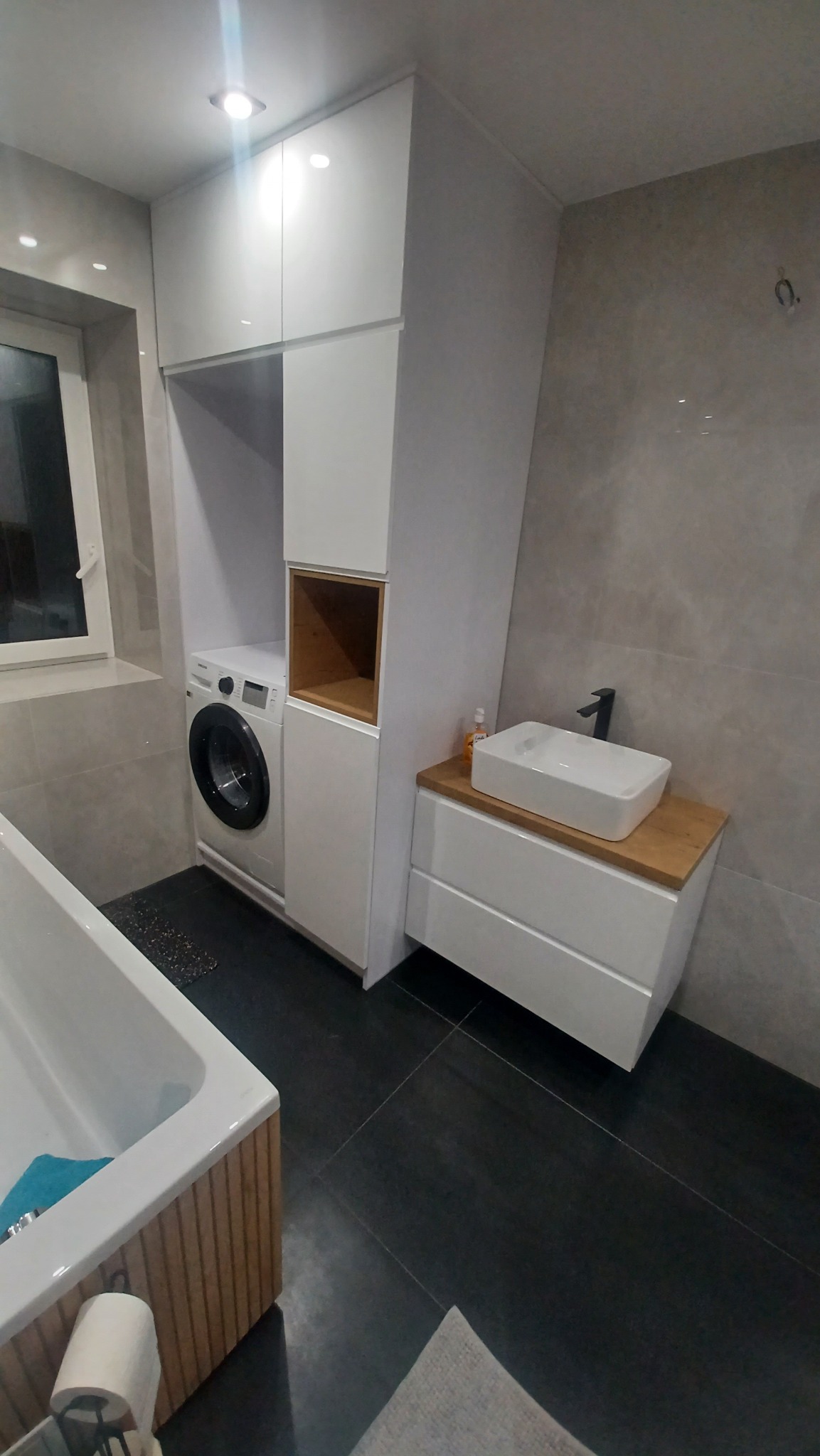 Biała zabudowa łazienkowa z pralką, szafkami i umywalką nablatową na drewnianym blacie, czarne płytki podłogowe, minimalistyczny styl.