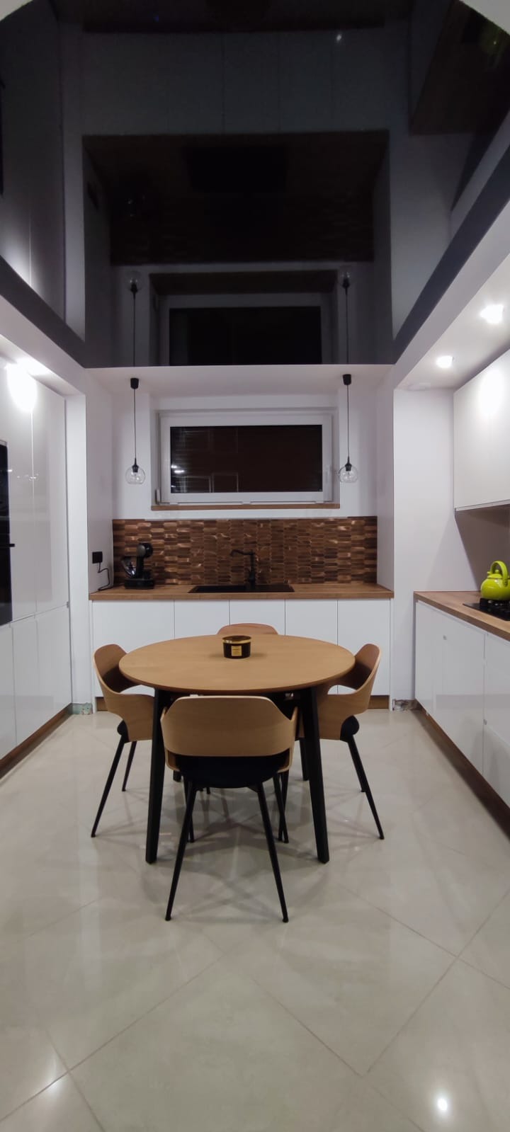 Nowoczesna kuchnia z białymi szafkami, drewnianym blatem i mozaiką, okrągły stół z czterema krzesłami w centrum, czarne, lśniące wykończenie sufitu.