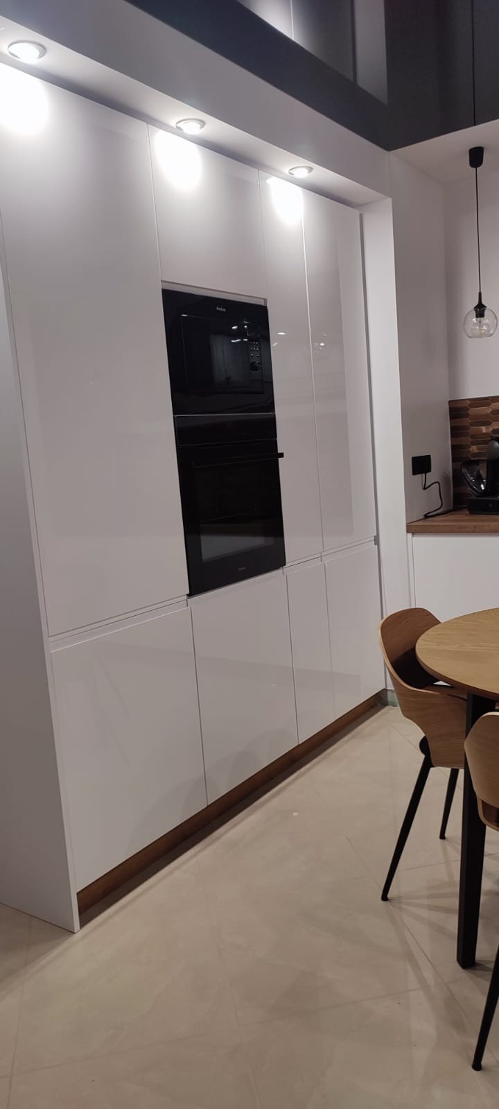 Biała, minimalistyczna zabudowa kuchenna z wbudowanym piekarnikiem i mikrofalówką Amica, widoczny fragment drewnianego stołu i krzesła w tle.