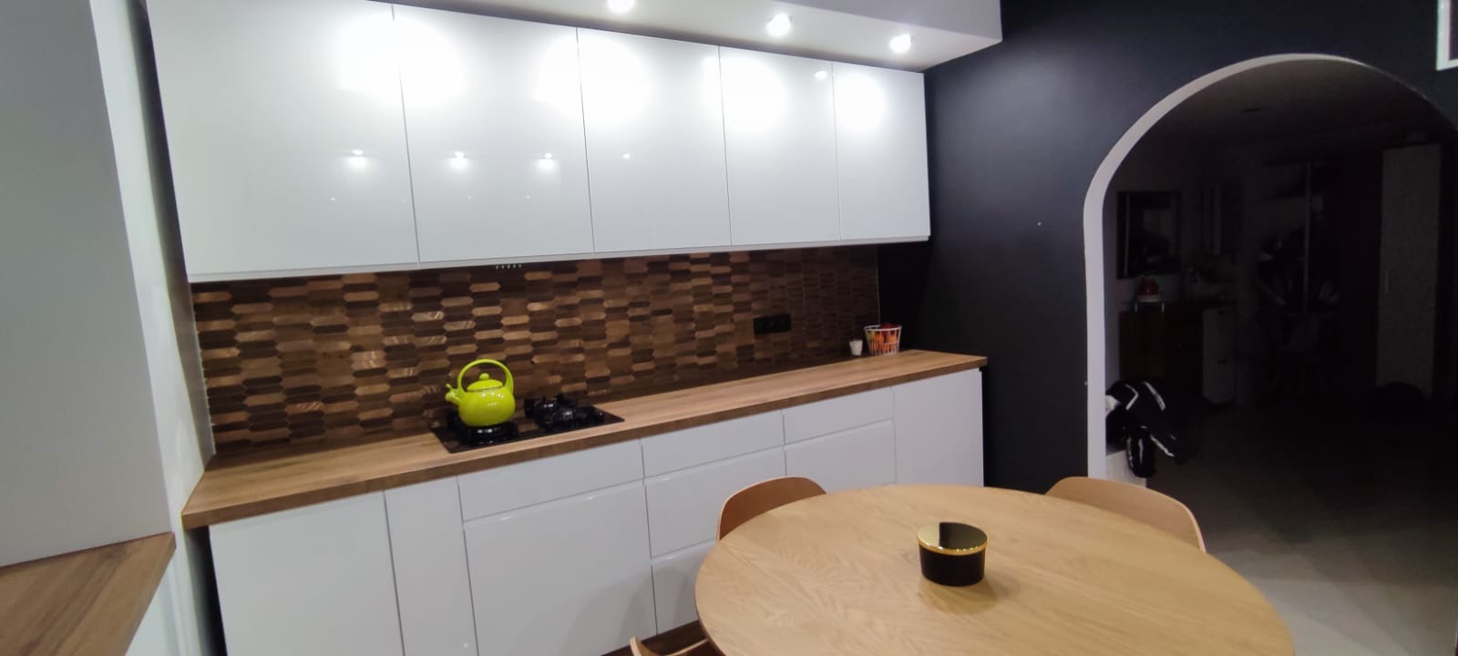 Nowoczesna kuchnia z białymi, błyszczącymi szafkami, drewnianym blatem i mozaiką na ścianie. Na blacie stoi zielony czajnik obok płyty indukcyjnej. W tle okrągły stół z drewna.