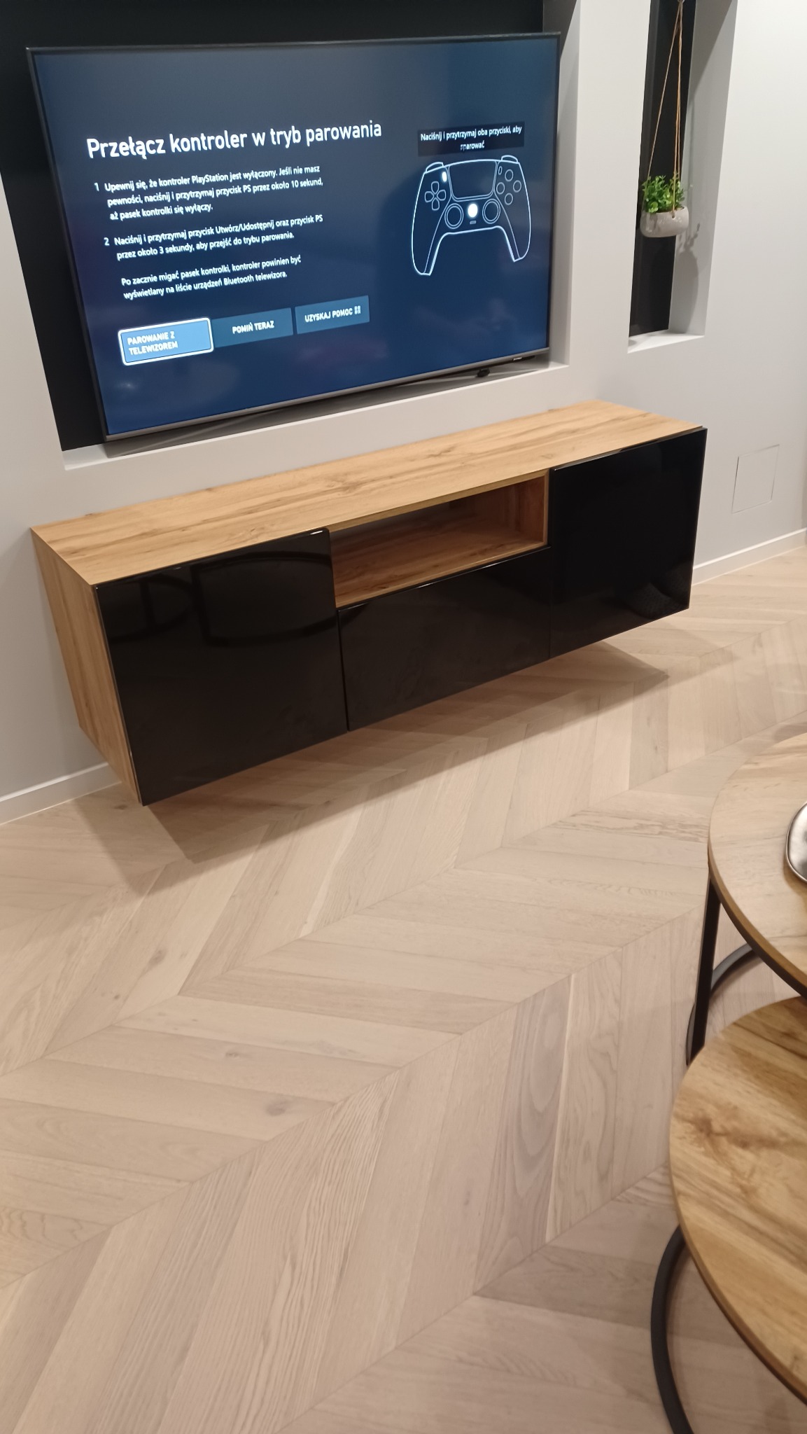 Nowoczesna szafka RTV wisząca z frontami w czarnym połysku i drewnianym blatem, pod telewizorem z instrukcją parowania kontrolera PlayStation, parkiet w jodełkę na podłodze.