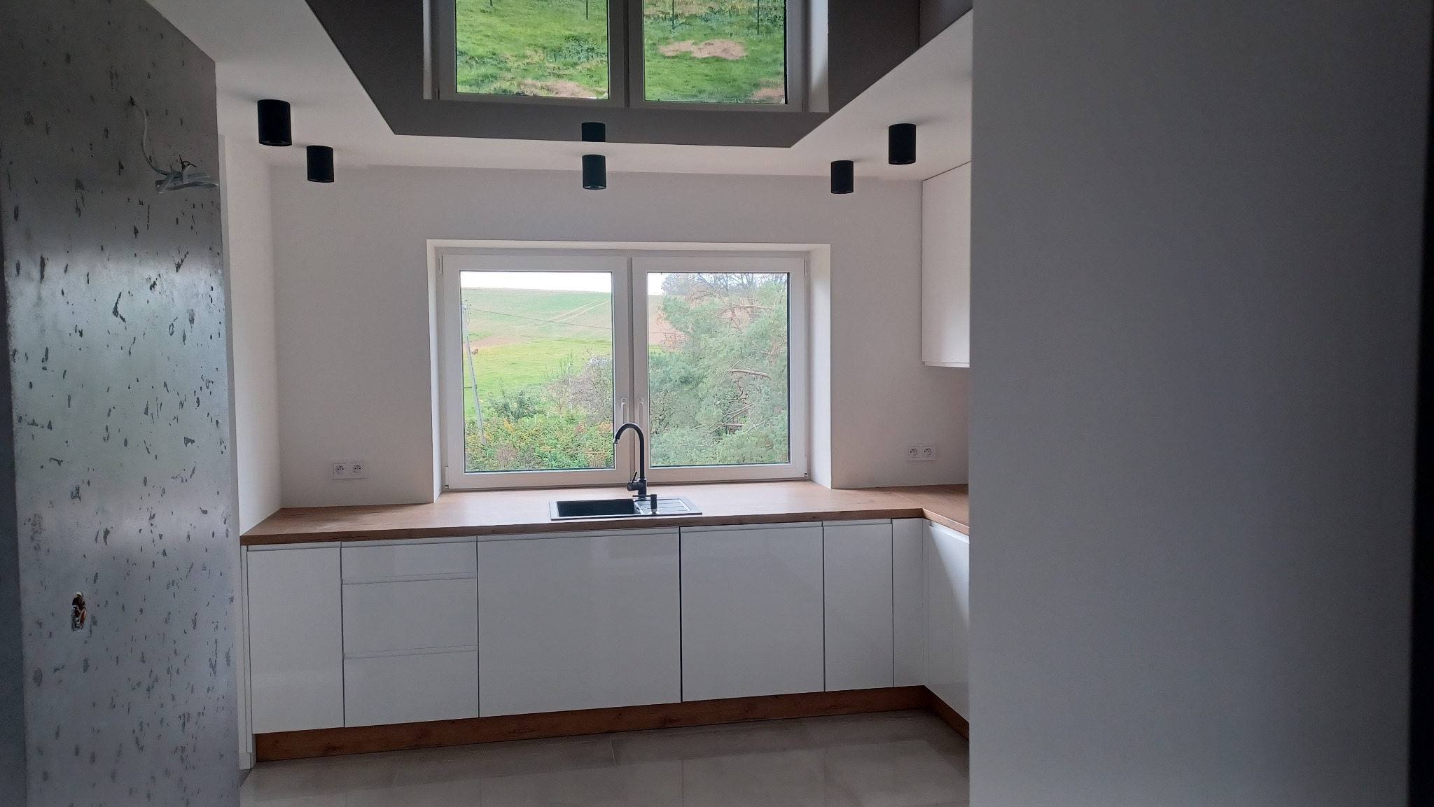 Nowoczesna kuchnia z białymi, błyszczącymi frontami szafek, drewnianym blatem i widokiem na zielony krajobraz za oknem