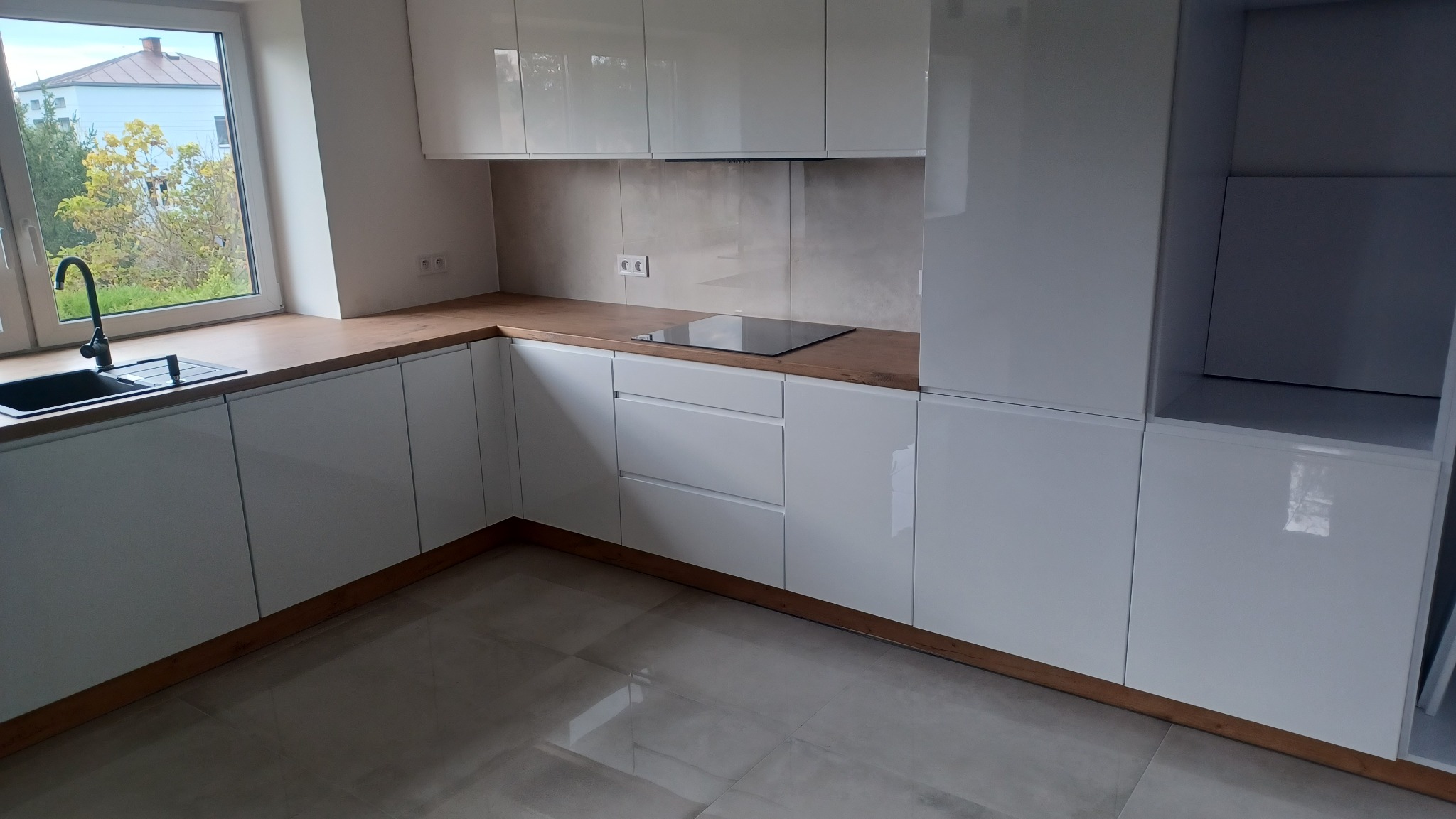 Nowoczesna, biała kuchnia z drewnianym blatem, czarnym zlewozmywakiem i płytą indukcyjną, widok z niskiego kąta, jasne płytki na podłodze.