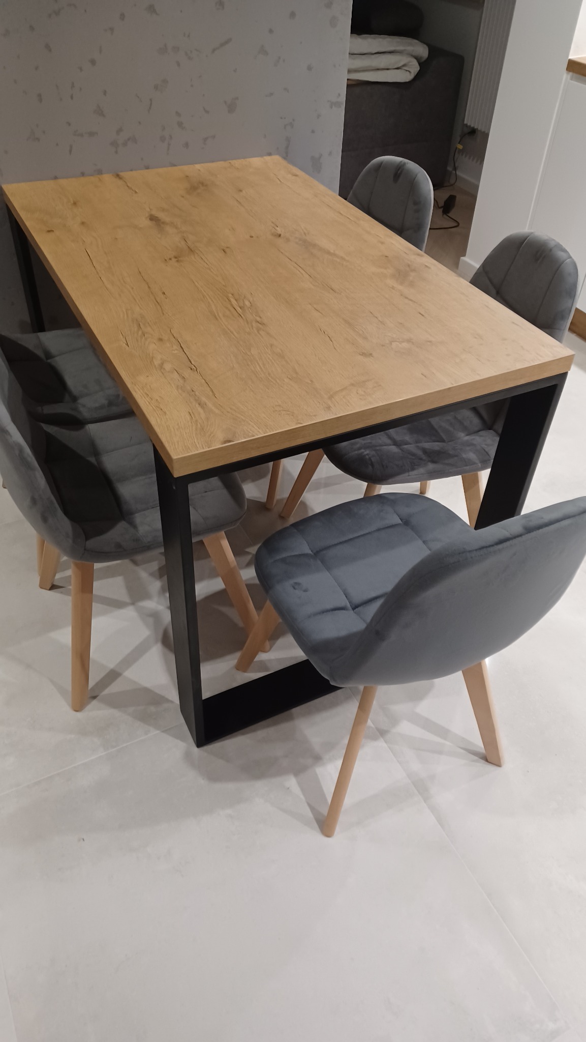 Stół z blatem imitującym drewno na czarnych nogach w stylu loftowym, otoczony czterema szarymi krzesłami tapicerowanymi z drewnianymi nogami, na jasnej podłodze.
