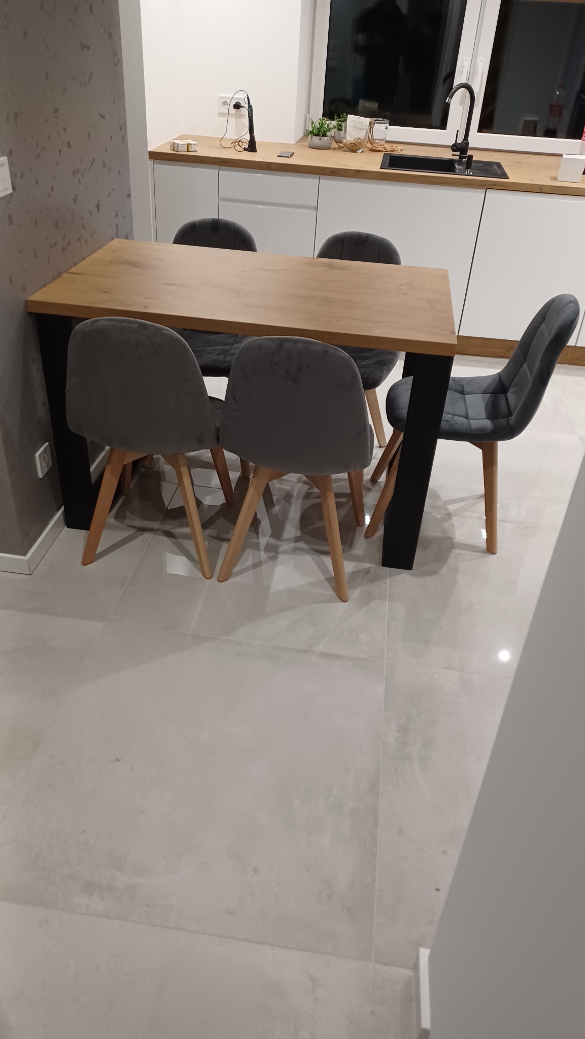 Drewniany stół z czarnymi nogami i szarymi krzesłami w nowoczesnej kuchni z białymi szafkami i szarą ścianą.