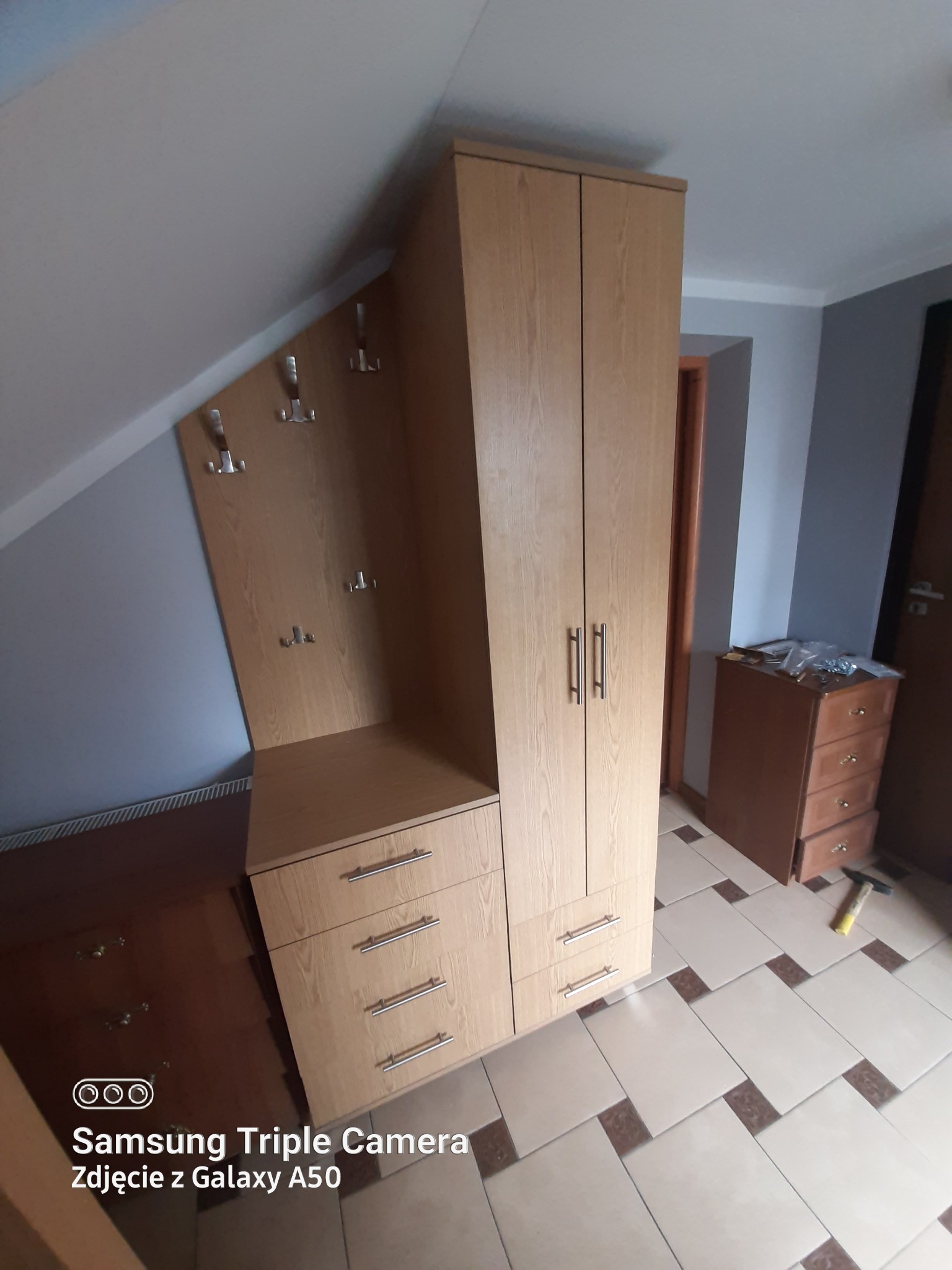 Jasna zabudowa meblowa w skosie poddasza, z szafą dwudrzwiową, komodą z szufladami i panelem z wieszakami, widoczny młotek na podłodze.